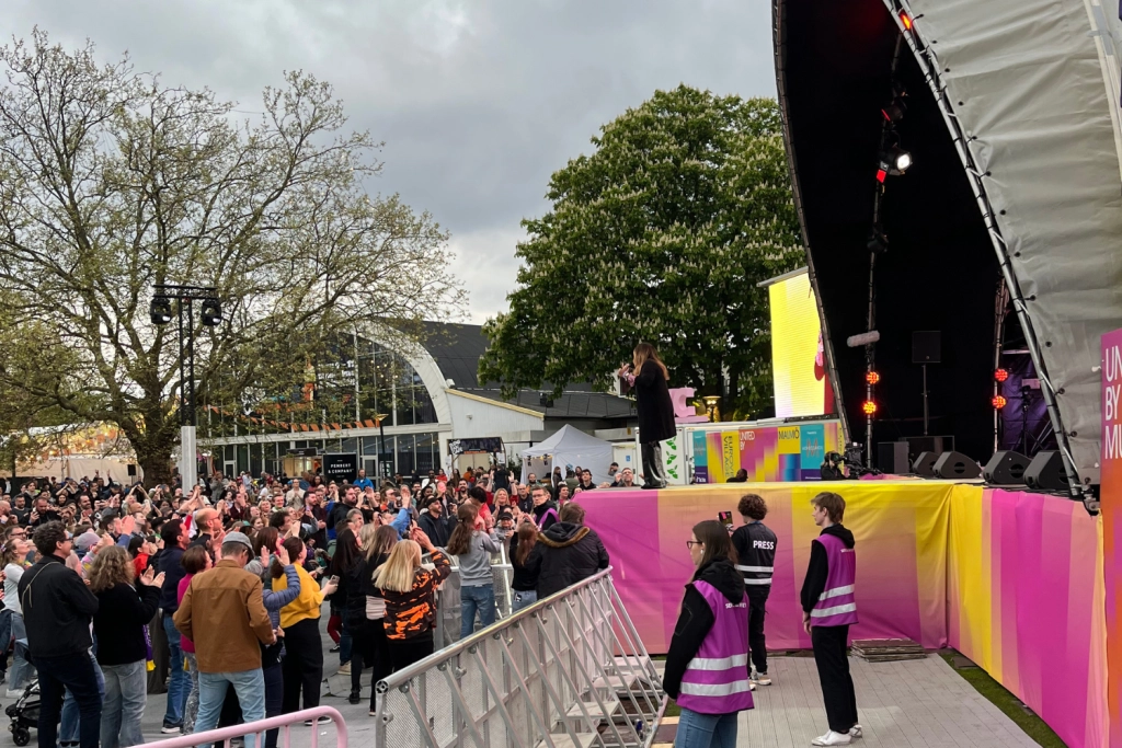 En artist på scen framför ett publikhav inne på Eurovision Village.